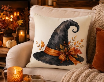 May include: A square throw pillow with an off-white background featuring a watercolor illustration of a black witch hat. The hat is adorned with an orange ribbon tied in a bow and decorated with autumn-colored leaves and small berries.