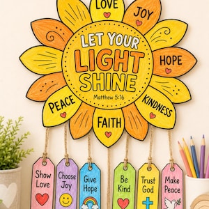 Op de afbeelding: Een zonnebloemvormige wandhanger met gele en oranje bloemblaadjes met de tekst "LET YOUR LIGHT SHINE" en "Matthew 5:16". Zes kleurrijke rechthoekige labels hangen eronder, elk voorzien van positieve zinnen zoals "Show Love" en "Be Kind".