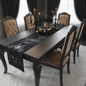 Può includere: Un tavolo da pranzo in legno scuro è apparecchiato per una cena a tema gotico. Un runner da tavolo nero con un disegno di teschio di coniglio è centrato sul tavolo. Candelabri neri con candele accese e teschi e uova decorative completano l'atmosfera inquietante. Sedie imbottite beige circondano il tavolo.