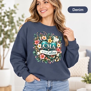 May include: A denim-coloured crewneck sweatshirt featuring a graphic print of a teal heart with a stethoscope, medical clipboard, and colourful flowers. The text "CMA is my Calling" is printed across the heart in white and dark blue lettering.