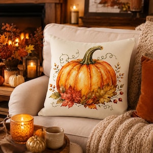 May include: A square throw pillow with an off-white background featuring a central illustration of a large orange pumpkin with a green stem, surrounded by detailed autumn leaves, small red berries, and delicate swirling vine tendrils.