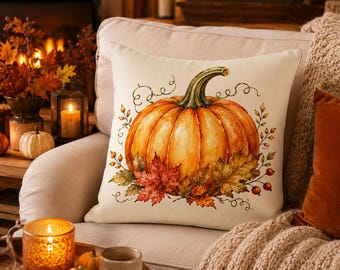 May include: A square throw pillow with an off-white background featuring a central illustration of a large orange pumpkin with a green stem, surrounded by detailed autumn leaves, small red berries, and delicate swirling vine tendrils.