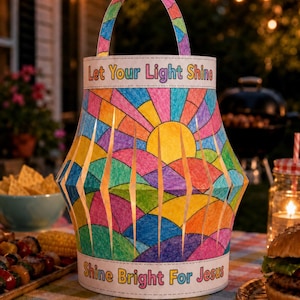 May include: A cylindrical paper lantern featuring a colorful stained-glass style pattern of a sun and rolling hills. The top band reads Let Your Light Shine and the bottom band reads Shine Bright For Jesus. It includes a matching paper handle.