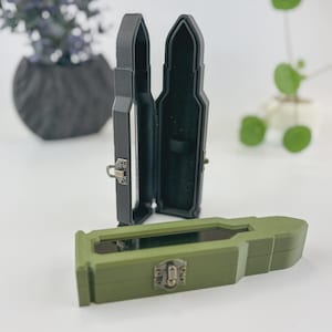 Könnte beinhalten: Schwarze und olivgrüne Schmuckkästchen in Form von Patronen mit Metallverschlüssen. Eine schwarze Box ist geöffnet und zeigt einen Samtinnenraum. Die olivgrüne Box ist geschlossen. Die Boxen stehen auf einer weißen Oberfläche.