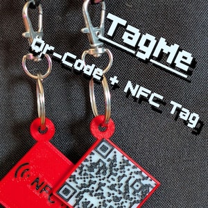 Puede incluir: Dos etiquetas NFC rojas en forma de diamante con llaveros y cierres plateados. Una etiqueta muestra un código QR, la otra un símbolo NFC. El texto "TagMe" y "qr-code + NFC tag" están en la imagen.