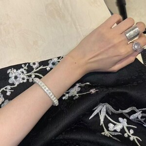 Op de afbeelding: Een sprankelende diamanten armband siert een pols, aangevuld met twee ringen. De ene ring is een brede, gegraveerde band, de andere heeft een grote, ronde edelsteen. De sieraden zijn geplaatst op een zwarte stof met witte bloemenborduurwerk.
