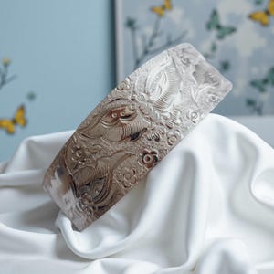Puede incluir: Un brazalete plateado con un diseño en relieve de flores y pájaros. El brazalete ancho se exhibe sobre una superficie de satén blanco, con un fondo azul claro y detalles de mariposas.