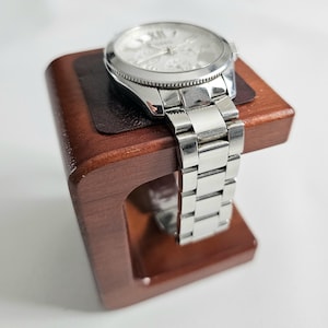 May include: A silver wristwatch with a white face and Roman numerals sits on a dark brown wooden watch stand. The watch features a stainless steel strap and a round face. The stand is a rich, dark wood colour.