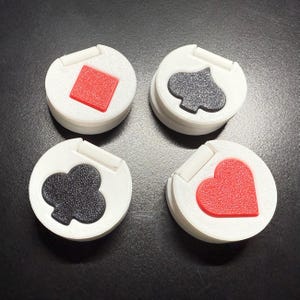 Può includere: Quattro gettoni di gioco circolari bianchi con simboli di carte da gioco: un rombo rosso, un picche nero, un trifoglio nero e un cuore rosso. Ogni pezzo ha una piccola linguetta bianca incernierata.