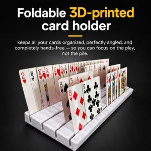 May include: A light grey, 3D-printed card holder featuring multiple parallel slots designed to hold playing cards upright. The text "Foldable 3D-printed card holder" and "keeps all your cards organised, perfectly angled, and completely hands-free" is visible.