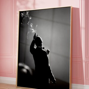 Pode incluir: Uma impressão de arte de parede em preto e branco emoldurada, apresentando a silhueta de uma pessoa de smoking em frente a um microfone. A figura segura um cigarro aceso, com volutas de fumaça subindo contra um fundo escuro e contrastante.