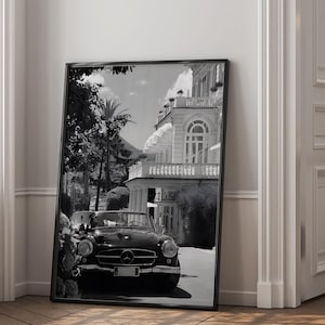 Puede incluir: Impresión en blanco y negro enmarcada que presenta un coche descapotable clásico estacionado frente a un gran edificio. La imagen captura una estética vintage con detalles arquitectónicos y exuberante vegetación, evocando lujo y viajes.
