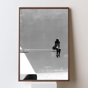 May include: A framed black and white photograph depicting a silhouette of a person sitting on the edge of a diving board. The scene is set against a bright, hazy sky and water, with the diving board extending horizontally from a white structure.