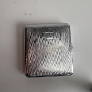 May include: A silver rectangular cigarette case with a textured, cross-hatched pattern. The case has a small rectangular emblem in the center and vertical lines. The case is closed with a small clasp.