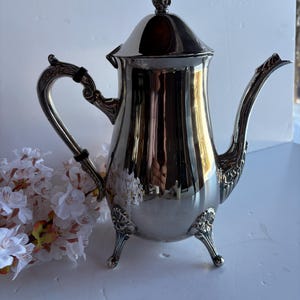 Puede incluir: Una cafetera plateada con un pico y asa curvos. La cafetera tiene una tapa decorativa y se apoya en tres patas ornamentadas. El asa tiene una banda negra. La superficie de la cafetera es muy reflectante.