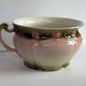 May include: A vintage ceramic chamber pot with a handle. The pot features a pink and green color scheme with decorative floral accents. The interior is a cream color.