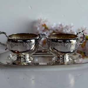 Puede incluir: Un juego de té plateado con dos tazas con asas y una bandeja rectangular. Las tazas y la bandeja tienen detalles ornamentados, incluido un diseño de vid. El juego está colocado sobre una superficie blanca con flores borrosas en el fondo.