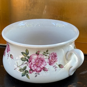 May include: White ceramic bowl with a handle, adorned with pink rose floral patterns and green leaves. The bowl features a wide rim and a curved handle. The background is a gold colour.