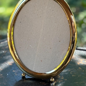 May include: A gold-colored, oval-shaped picture frame. The frame has a shiny, reflective surface and is supported by three small, round feet. The interior of the frame is a neutral gray color, ready for a photograph or artwork.