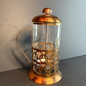 Op de afbeelding: Een cafetière met een heldere glazen kan en een koperkleurig metalen frame. Het frame heeft een decoratief cirkelvormig patroon. Het deksel en de handgreep zijn ook koperkleurig. De cafetière is ontworpen voor het zetten van koffie.