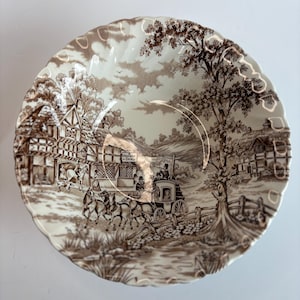 May include: A decorative ceramic plate with a brown transferware design. The plate features a rural landscape scene with a horse-drawn carriage, buildings, and trees. The rim has a scalloped edge.