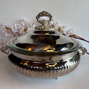 Könnte beinhalten: Eine silberfarbene Servierschüssel mit Glasdeckel und Ziergriff. Die Schüssel hat ein geripptes Design und dekorative Griffe. Der Deckel ist mit einem detaillierten Griff versehen. Ideal zum Servieren von Speisen.