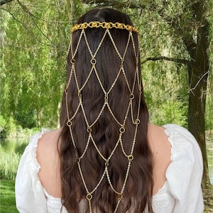 May include: A gold-toned metal hair accessory featuring a circular headband connected to a cascading lattice of fine chains and small rings that drape down the back. The structure creates a diamond-shaped pattern across the hair surface.