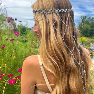K&ouml;nnte beinhalten: Ein silberfarbener Ketten-Kopfschmuck mit einem horizontalen Band aus ineinandergreifenden Metallringen, das auf der Stirn aufliegt, sowie mehreren drapierten Kettenstr&auml;ngen, die nach hinten verlaufen und einen kaskadierenden Netzeffekt bilden.