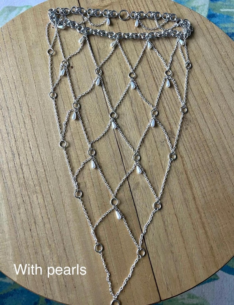 K&ouml;nnte beinhalten: Ein silberfarbenes Kettenhemd-Handaccessoire mit einem rautenf&ouml;rmigen Gittermuster aus miteinander verbundenen Metallringen und feinen Ketten. Kleine, wei&szlig;e, tropfenf&ouml;rmige Kunstperlen h&auml;ngen an den Verbindungsstellen der Kettenglieder.