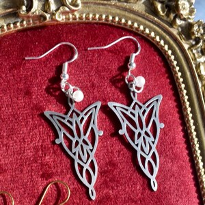 K&ouml;nnte beinhalten: Ein Paar silberfarbene Ohrh&auml;nger mit einem komplizierten, symmetrischen durchbrochenen Metalldesign und einer spitz zulaufenden Unterseite. Jeder Ohrring h&auml;ngt an einem silberfarbenen Haken und ist mit einer kleinen, runden wei&szlig;en Perle versehen.