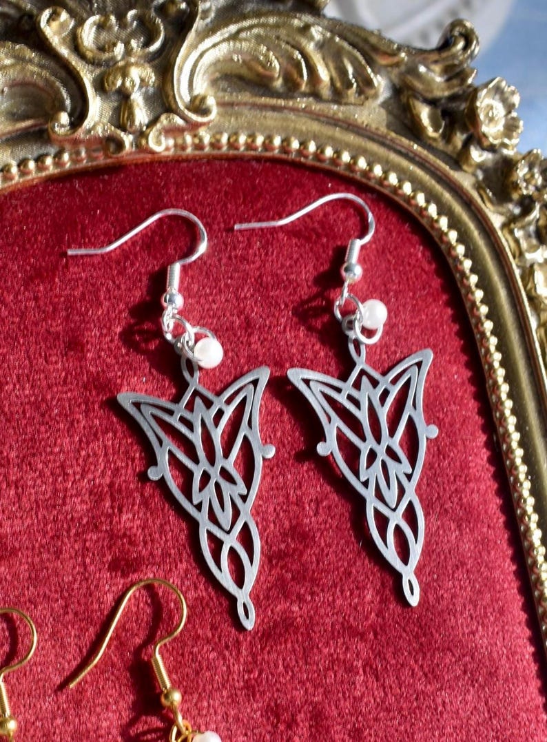 K&ouml;nnte beinhalten: Ein Paar silberfarbene Ohrh&auml;nger mit einem komplizierten, symmetrischen durchbrochenen Metalldesign und einer spitz zulaufenden Unterseite. Jeder Ohrring h&auml;ngt an einem silberfarbenen Haken und ist mit einer kleinen, runden wei&szlig;en Perle versehen.
