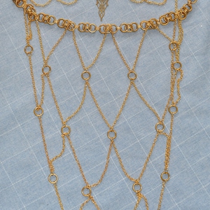 May include: A gold-toned chain mail accessory featuring a top band of interlocking circular rings, a central filigree pendant, and a cascading web of fine link chains connected by smaller rings, creating a structured, draped, net-like appearance.