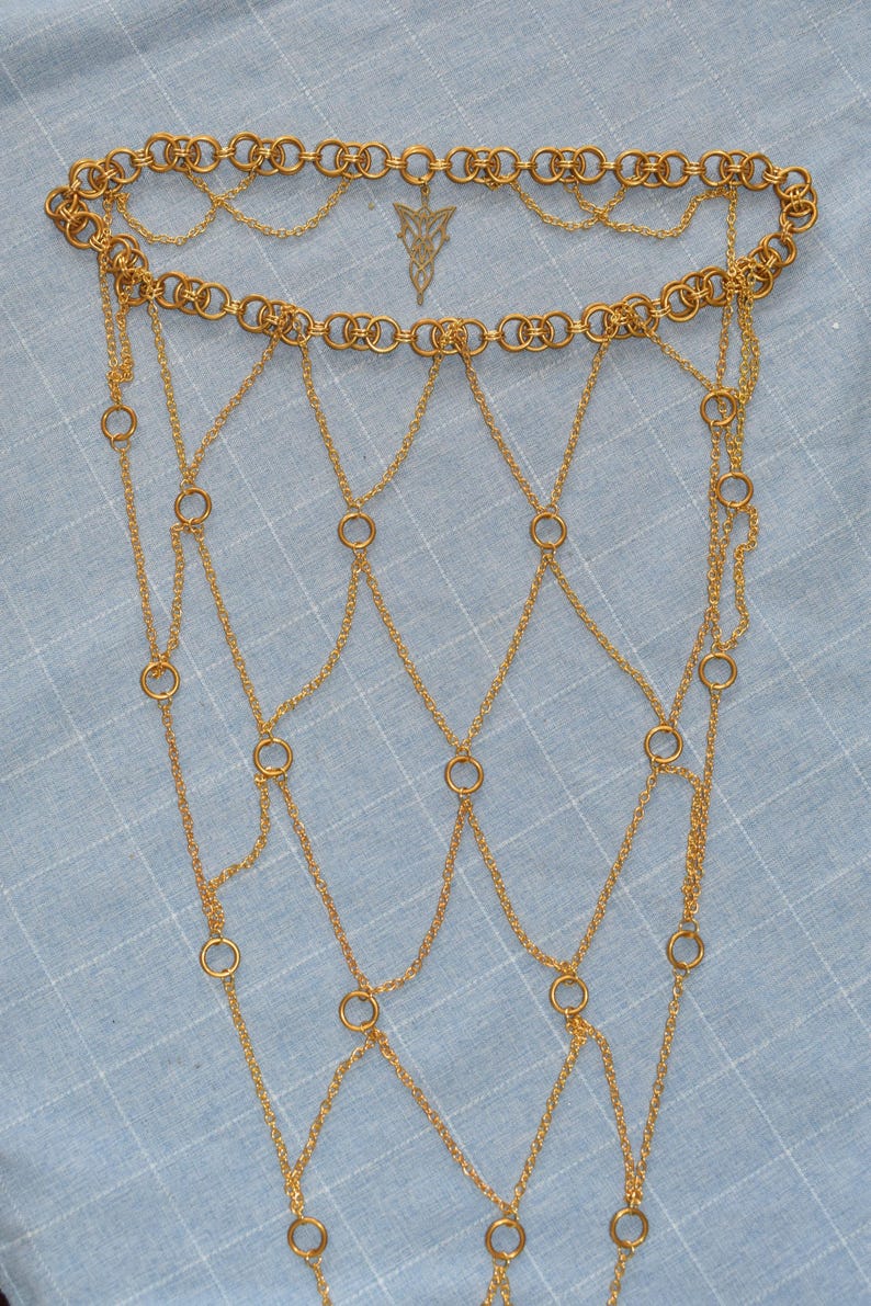 May include: A gold-toned chain mail accessory featuring a top band of interlocking circular rings, a central filigree pendant, and a cascading web of fine link chains connected by smaller rings, creating a structured, draped, net-like appearance.