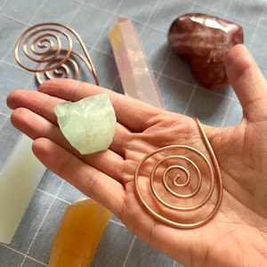 May include: A collection of crystals and copper wire art. The crystals include a light green, rough-cut stone, a clear quartz point with pink and yellow hues, and a reddish-brown heart-shaped stone. Two copper wire spirals are also visible.