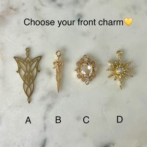 May include: Four gold-toned metal charms labeled A, B, C, and D. Charm A is a filigree leaf shape, B is a sword, C is an oval frame with a clear stone, and D is a starburst with a central stone. The text reads: Choose your front charm.