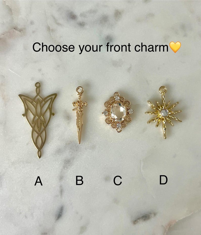 May include: Four gold-toned metal charms labeled A, B, C, and D. Charm A is a filigree leaf shape, B is a sword, C is an oval frame with a clear stone, and D is a starburst with a central stone. The text reads: Choose your front charm.