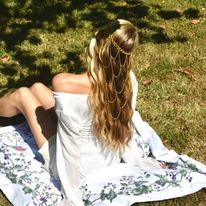May include: A gold-toned metal headpiece featuring multiple draped chains that hang down from a circular band. The headpiece is worn over long blonde hair, paired with a white long-sleeved top and a white fabric featuring a botanical print.