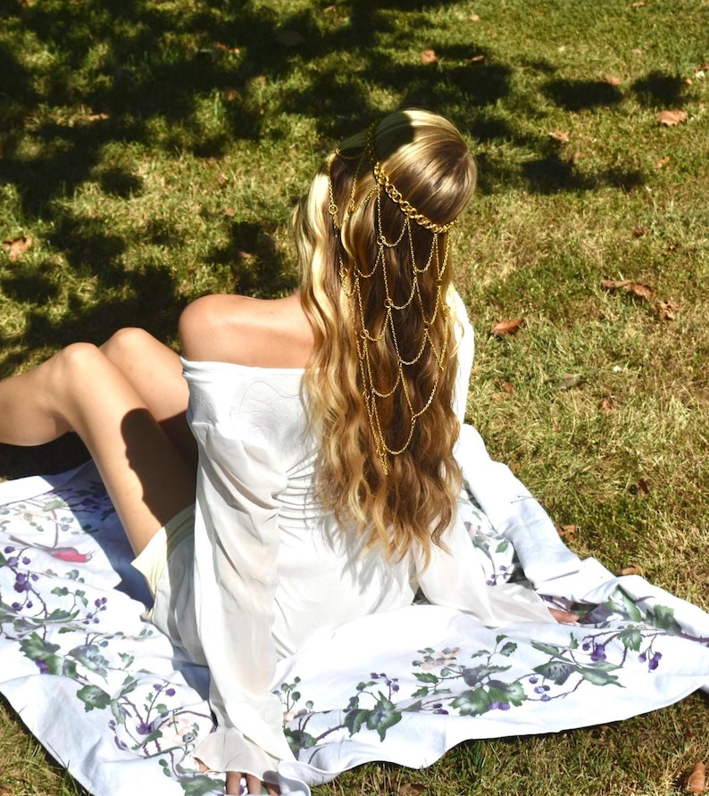May include: A gold-toned metal headpiece featuring multiple draped chains that hang down from a circular band. The headpiece is worn over long blonde hair, paired with a white long-sleeved top and a white fabric featuring a botanical print.