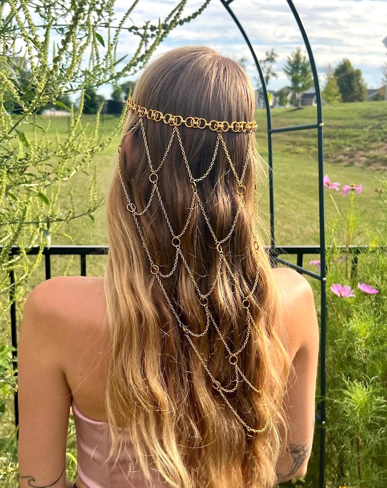 May include: A gold-toned headpiece featuring a horizontal band of interlocking circular links. Multiple delicate gold chains drape downward from the band, connected by small circular rings, creating a layered, cascading net-like effect.