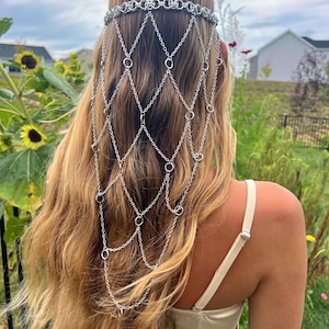 K&ouml;nnte beinhalten: Ein silberfarbenes Haaraccessoire mit einem Stirnband im Kettenhemd-Stil, das mit einem kaskadierenden Netz aus Metallgliedern und Ringen verbunden ist, die &uuml;ber den Hinterkopf fallen. Das St&uuml;ck besteht aus miteinander verbundenen silbernen Metallketten.