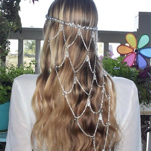 K&ouml;nnte beinhalten: Ein silberfarbenes Haaraccessoire mit einem zarten Ketten-Kopfschmuck, bestehend aus mehreren drapierten Str&auml;ngen aus klaren Kristallen und kleinen wei&szlig;en Perlen. Das Design umfasst ein horizontales Band am Oberkopf mit kaskadierenden Ketten.