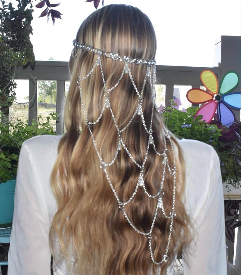 K&ouml;nnte beinhalten: Ein silberfarbenes Haaraccessoire mit einem zarten Ketten-Kopfschmuck, bestehend aus mehreren drapierten Str&auml;ngen aus klaren Kristallen und kleinen wei&szlig;en Perlen. Das Design umfasst ein horizontales Band am Oberkopf mit kaskadierenden Ketten.