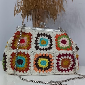 May include: A handmade crochet clutch with a silver metal clasp and chain strap. The bag features a patchwork design of colorful squares in shades of orange, yellow, green, brown, and blue, set against a cream background. A stylish accessory.