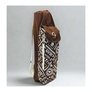 May include: A brown yoga mat bag with a drawstring closure and a side pocket. The bag features a white geometric pattern on the front and a cream-colored zipper. The bag has a shoulder strap and the text "O2" on the side pocket.