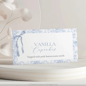 May include: A white card with blue floral border and text that reads "Vanilla Cupcakes". The card is decorated with a blue ribbon and the text "Topped with pink buttercream swirls". The card is on a white pedestal.