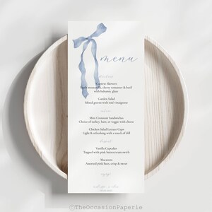 May include: A menu card featuring a watercolour blue ribbon design and handwritten text. The menu lists starters, salads, main courses, and desserts. The card is placed on a light wooden plate.