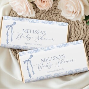 Könnte beinhalten: Zwei rechteckige Babyparty-Gastgeschenke mit weißem Hintergrund und blauem Blumenrand. Der Text "MELISSA'S Baby Shower" ist in eleganter Schrift dargestellt, darunter das Datum. Eine blaue Schleife befindet sich auf der linken Seite.