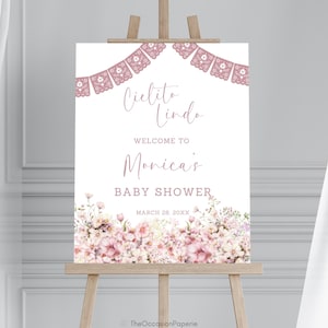 Op de afbeelding: Een babyborrelbord op een houten ezel. Het bord is wit met roze bloemaccenten en de tekst "Cielito Lindo", "Welkom bij Monica's Baby Shower" en de datum. Een banner van roze papieren uitsparingen staat bovenaan.