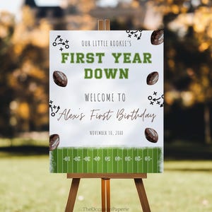May include: A white sign on a wooden easel with the text "OUR LITTLE ROOKIE'S FIRST YEAR DOWN" in green and black. The sign welcomes guests to "Alex's First Birthday" and includes football graphics and a field design at the bottom.