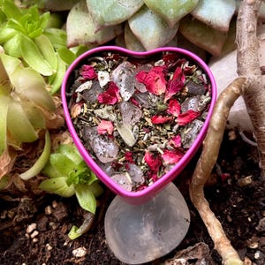 May include: A pink heart-shaped container filled with dried herbs, rose petals, and clear crystals. The container rests on a large, smooth, clear quartz crystal. The background features green succulents and brown soil.